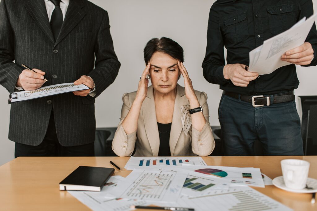 White businesswoman sits a conference table with papers of charts and graphs in front of her. She has her hands at her temples, looking stressed. She's flanked by two professionals, whose heads we don't see.
